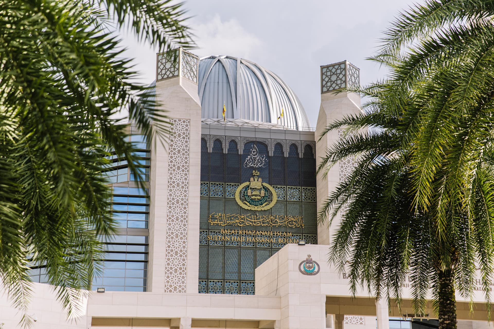 The dome and tiled façade of Balai Khazanah Islam Sultan Haji Hassanal Bolkiah, framed by palm fronds.